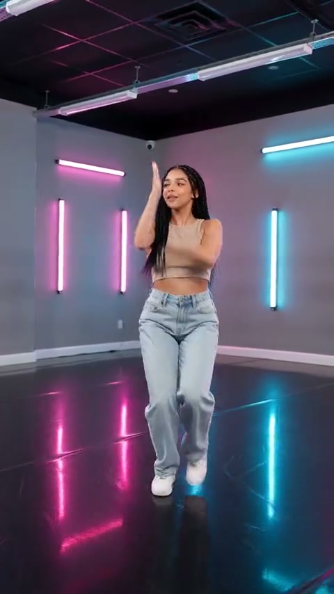 Beeld uit een externe video van een solodanser in een jeans en witte sportschoenen die één arm en één voet optilt in een roze-cyaan neonstudio